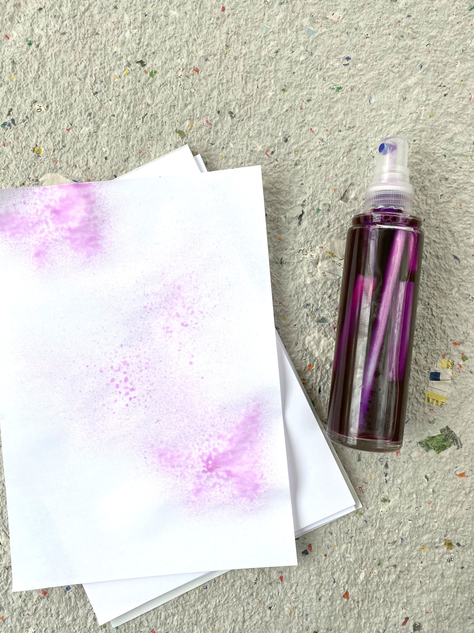 Neben einer Sprühflasche gefüllt mit Patronen aus Filzstiften und dadurch lila verfärbten Wasser liegt ein Blatt Papier mit einem gesprühten lila Farbklecks.