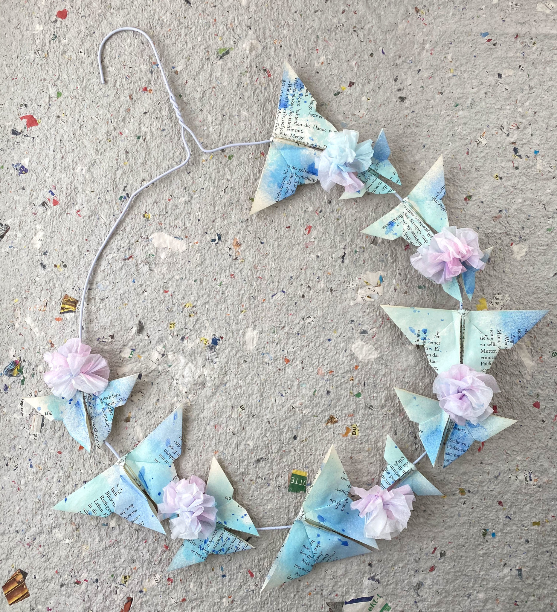Drahtbügel aus der Wäscherei zu einem Kreis gebogen. Rundherum sind Origami-Schmetterlinge aus alten Buchseiten geklebt. Die Schmetterlinge sind mit verschiedenen Blautönen besprüht. Für Auflockerung sorgen kleine rosa besprühte Blumen aus Kosmetiktüchern.